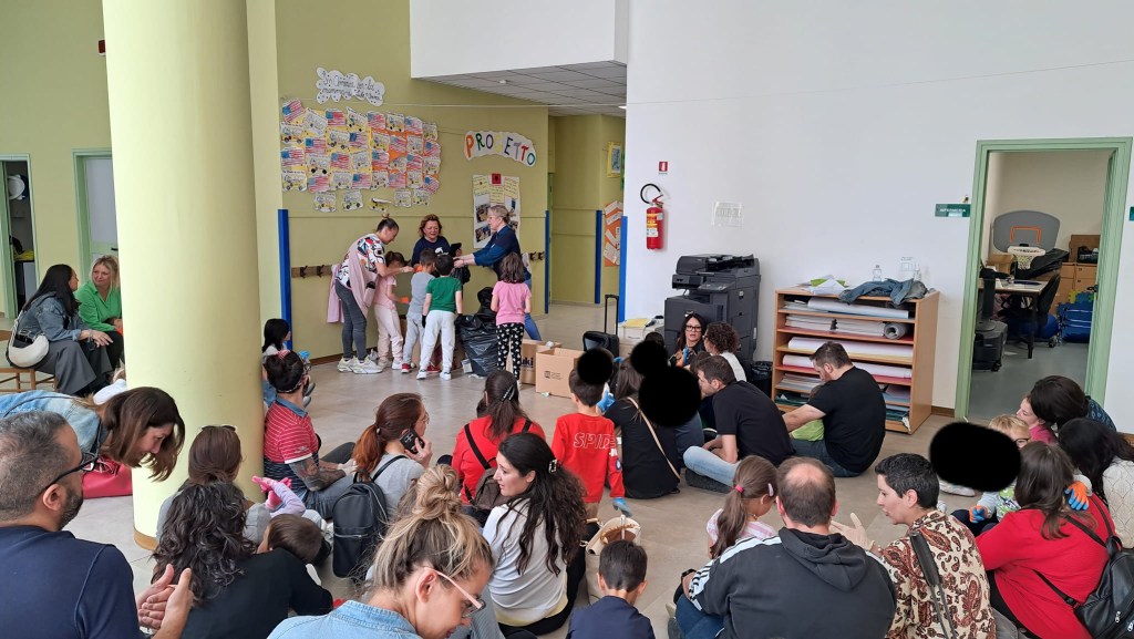 A group of children and parents participating in an environmental awareness event at the Il Girasole kindergarten in Corciano. The room is lively with adults and kids engaged in activities, while a banner related to the project hangs on the wall.