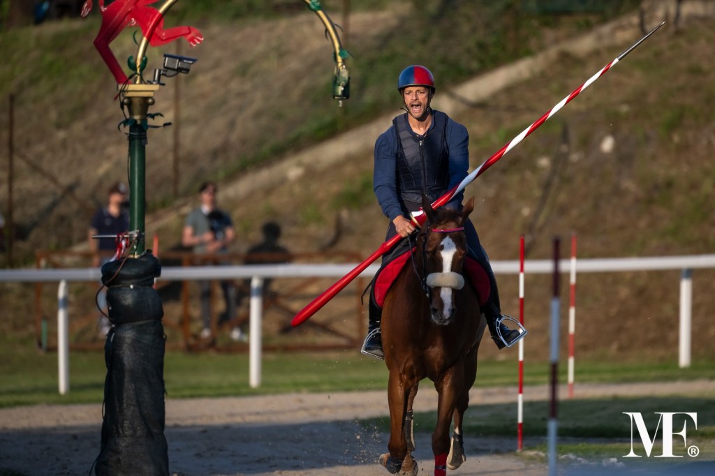Un cavaliere in sella a un cavallo, mentre manovra una lancia per afferrare un anello sospeso. Sullo sfondo, si possono vedere spettatori e un campo di gara.