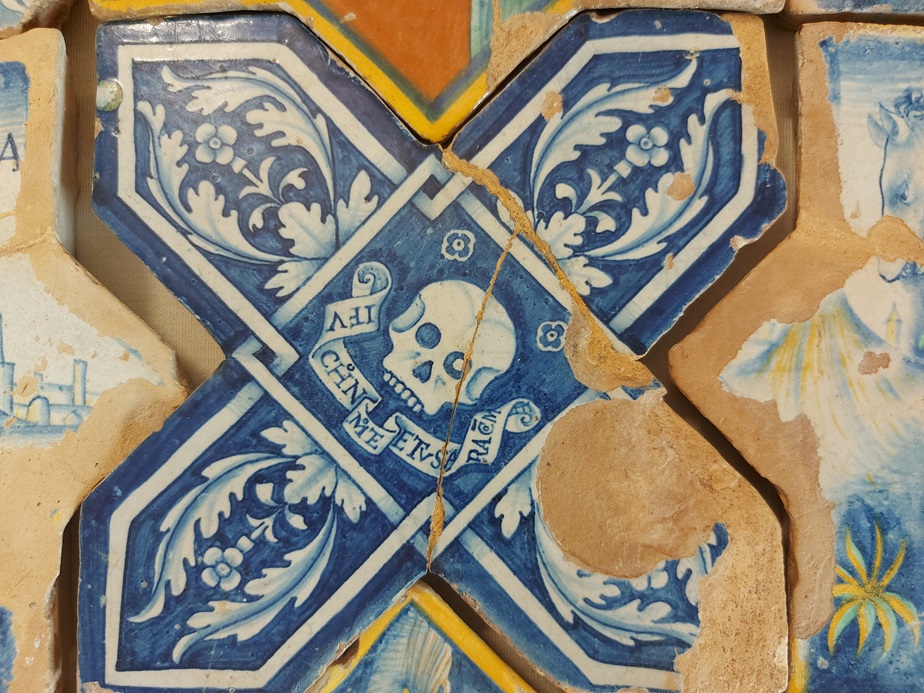 Dettaglio di una mattonella maiolicata con decorazioni blu e bianche, che include un teschio e motivi floreali, parte di un pavimento storico.