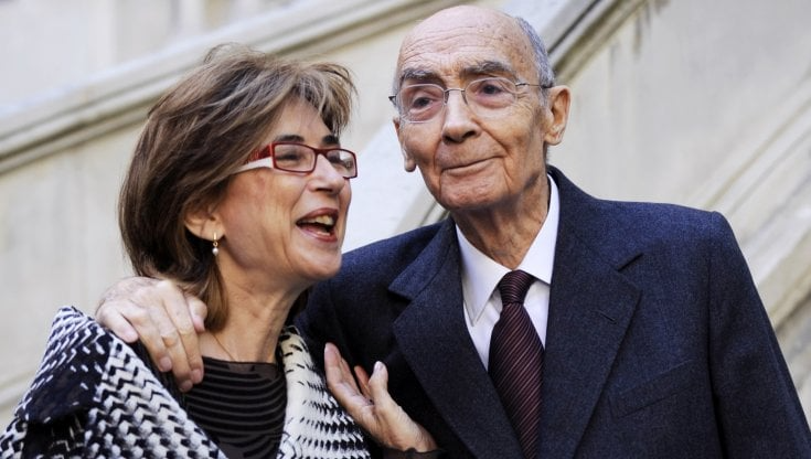 Due persone sorridenti in un ambiente esterno, uno dei quali indossa occhiali e un abito scuro, l'altro una sciarpa e un abito scuro, che sembrano conversare amichevolmente.