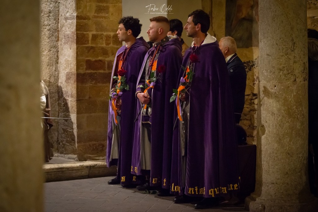 Tre cavalieri appartenenti al Terziere di Santa Maria in abiti di cerimonia, in attesa della benedizione, all'interno di una chiesa a Narni, con fiori in mano e un'atmosfera medievale.
