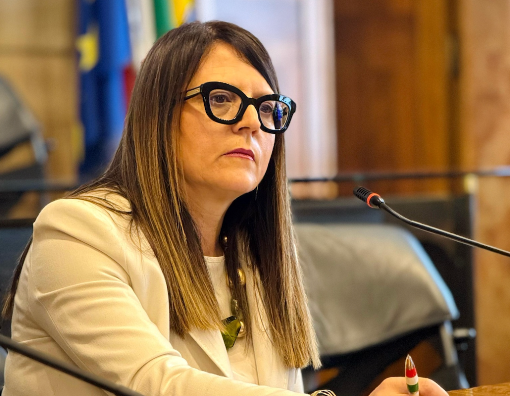 A woman with long hair and glasses sits at a desk in a formal setting, looking thoughtfully ahead. She wears a white blazer and has a decorative pen in her hand, with flags visible in the background.