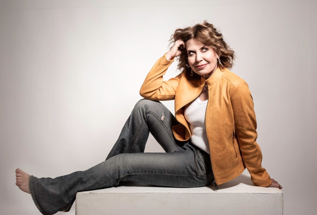 Sabina Guzzanti in posa su un supporto bianco, indossando una giacca in pelle gialla, una maglietta bianca e jeans. Sorridente, con i capelli mossi, trasmette un'atmosfera rilassata e creativa.