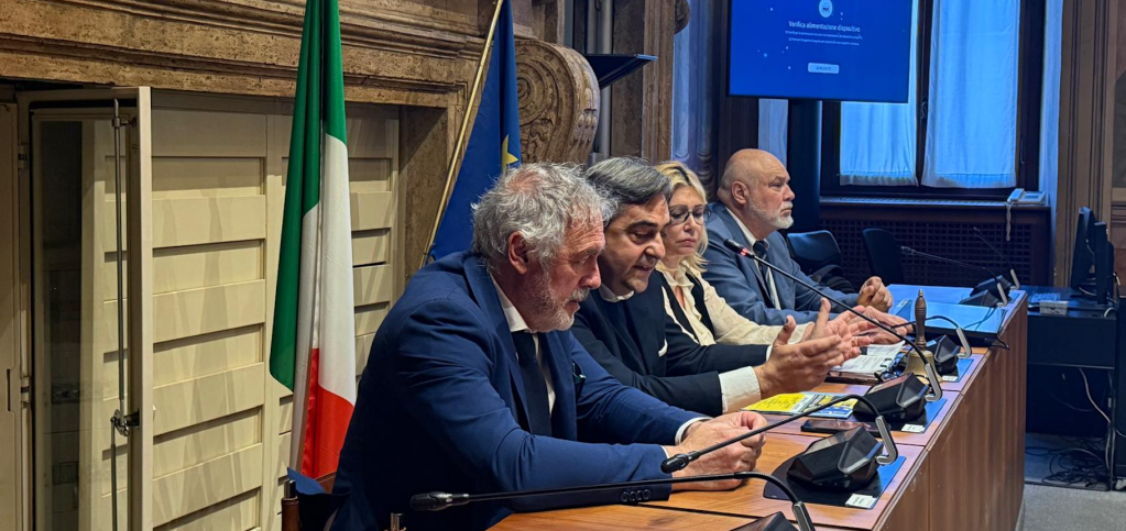 Un gruppo di relatori durante un convegno sulla NATO a Terni, con bandiere italiana ed europea sullo sfondo. I partecipanti discutono temi geopolitici ed economici.