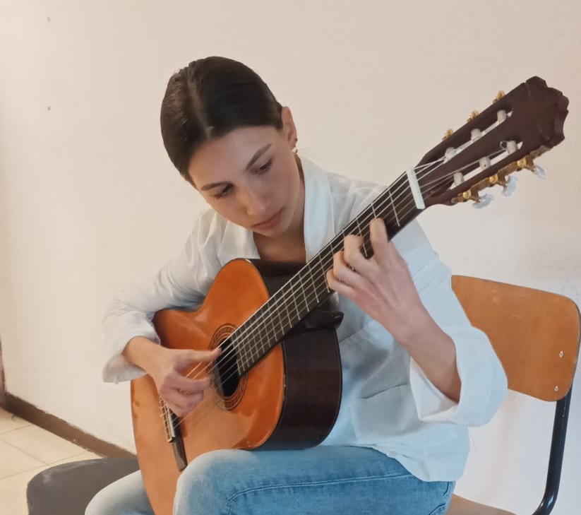 Una giovane donna suona la chitarra, concentrata, seduta su una sedia in una stanza con pareti chiare.