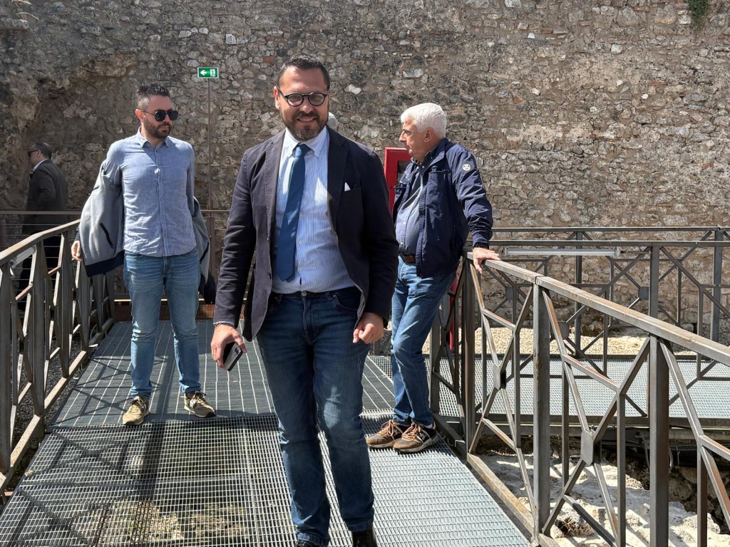 Three men walking on a metal walkway in front of a stone wall. One man is wearing glasses and a suit, while another is in casual attire with a shirt and jeans. The setting appears to be an archaeological site.
