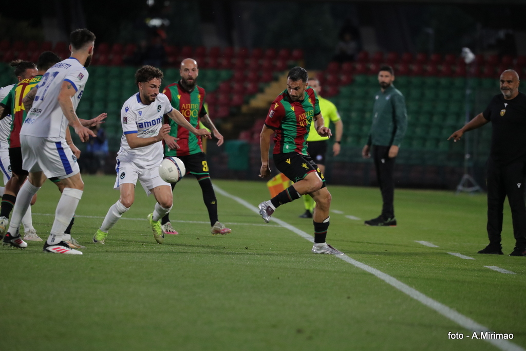 Un momento di gioco in una partita di calcio con un giocatore in maglia bianca che cerca di recuperare la palla da un avversario in maglia verde e rossa, mentre i compagni di squadra e l'arbitro osservano.