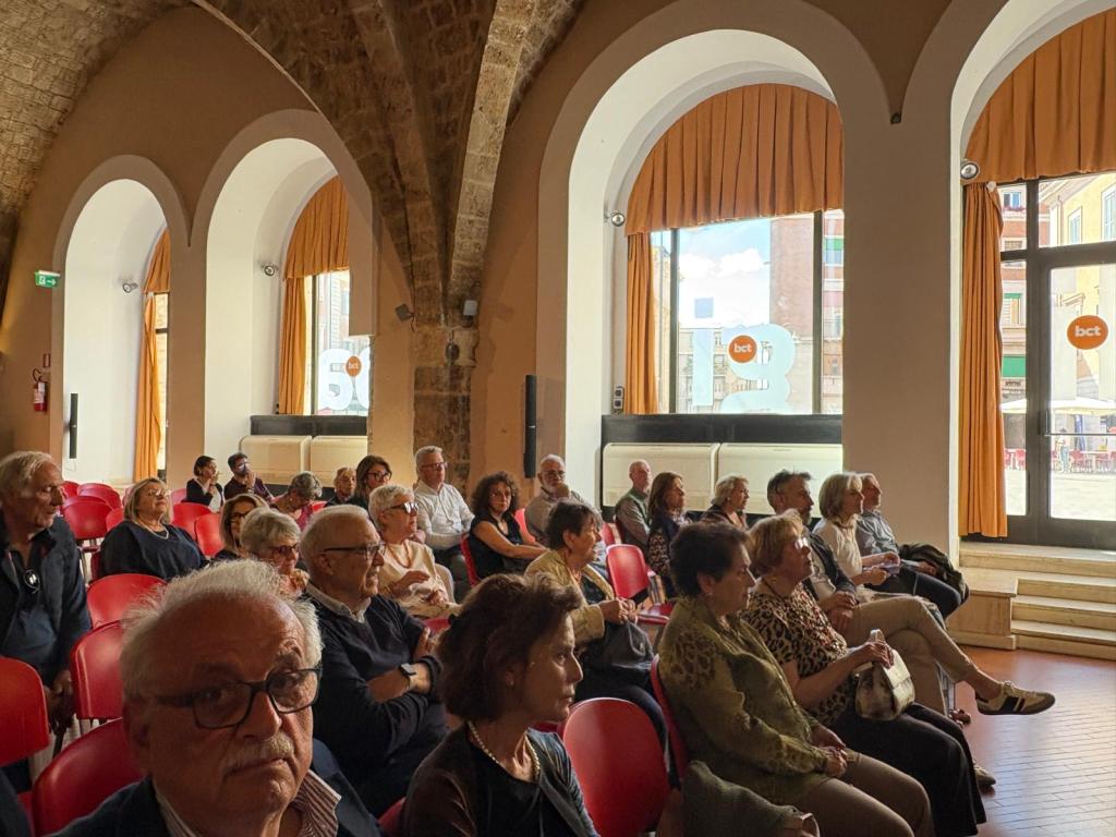 Un pubblico assorto durante la conferenza per la Giornata della Dante, con sedie rosse e finestroni che mostrano l'esterno della BCT.