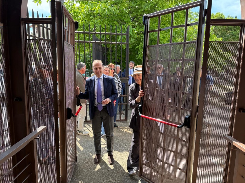 A group of people gathered at a gate, with two individuals actively opening the large gates. The scene is set outdoors with greenery in the background.