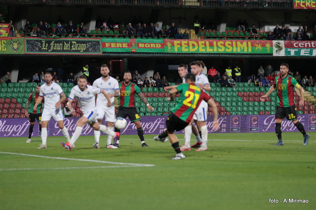 Giocatori in campo durante una partita di calcio, con la squadra in maglia rossa e verde in attacco e avversari in maglia bianca in difesa. Segnaposto pubblico e bandiere visibili sugli spalti.
