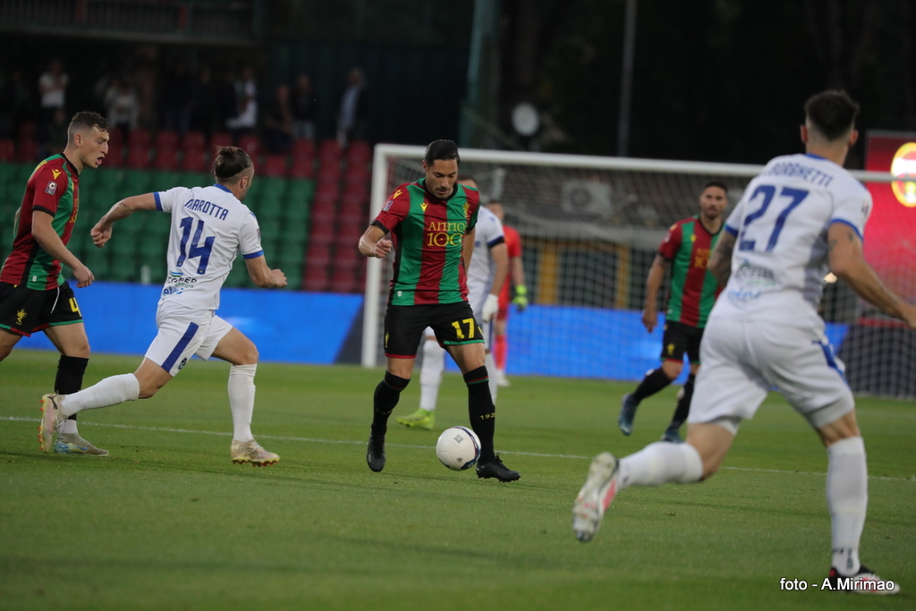 Giocatori in campo durante una partita di calcio, con un giocatore in uniforme con i colori rosso e verde che dribbla un avversario in maglia bianca.