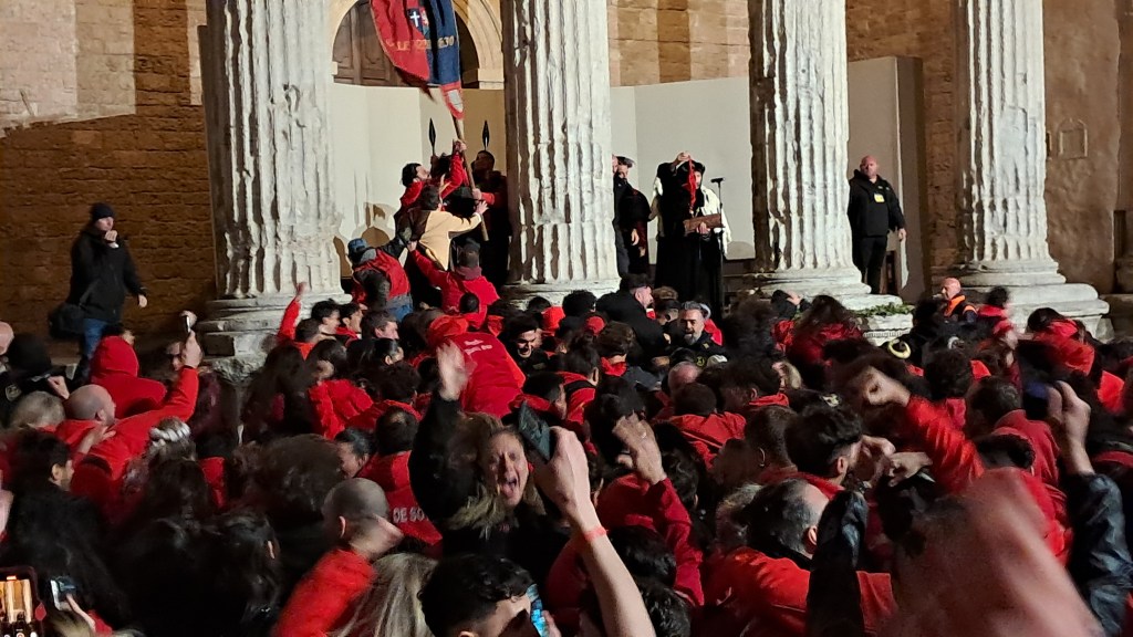 Folla vestita di rosso celebra la vittoria della Parte de Sotto al Calendimaggio di Assisi, con bandiere sollevate e festeggiamenti davanti a un palcoscenico.