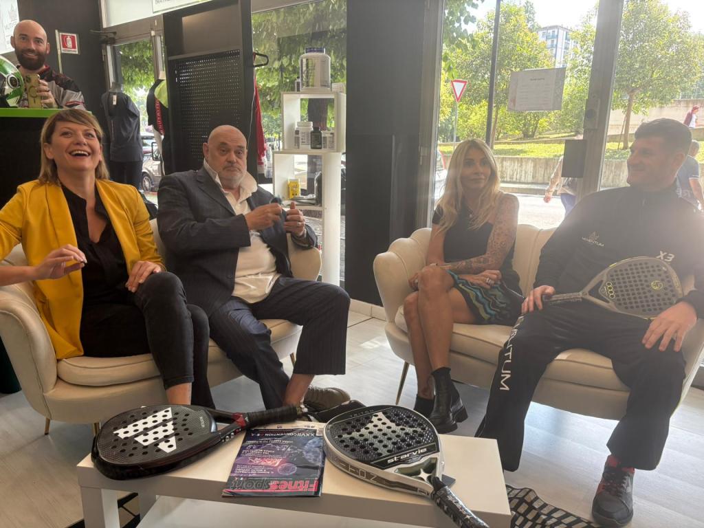 Quattro persone sedute su un divano in un ambiente luminoso, con racchette da padel e un giornale sulla tavola.