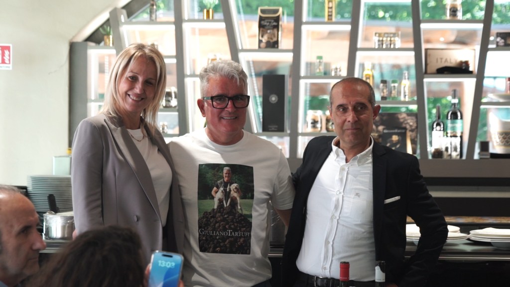 Tre persone sorridenti in una presentazione, con uno degli uomini indossando una maglietta con un'immagine di un cane e tartufi sullo sfondo.