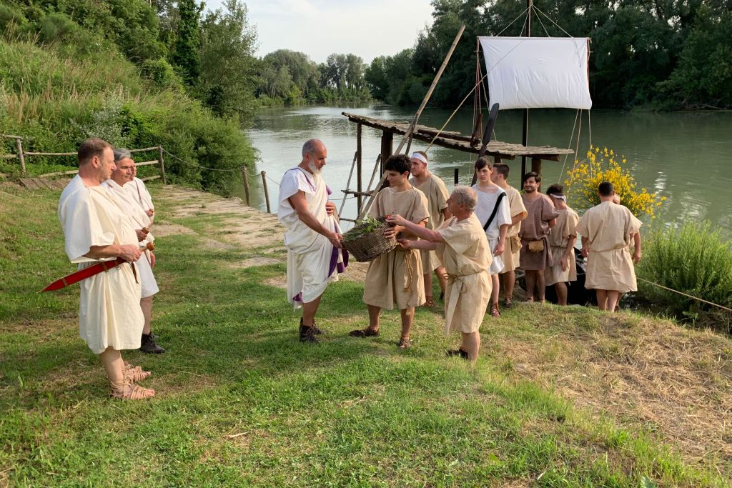 Gruppo di persone vestite in abiti romani tradizionali vicino a un fiume, interagendo tra di loro mentre un'imbarcazione è attraccata.