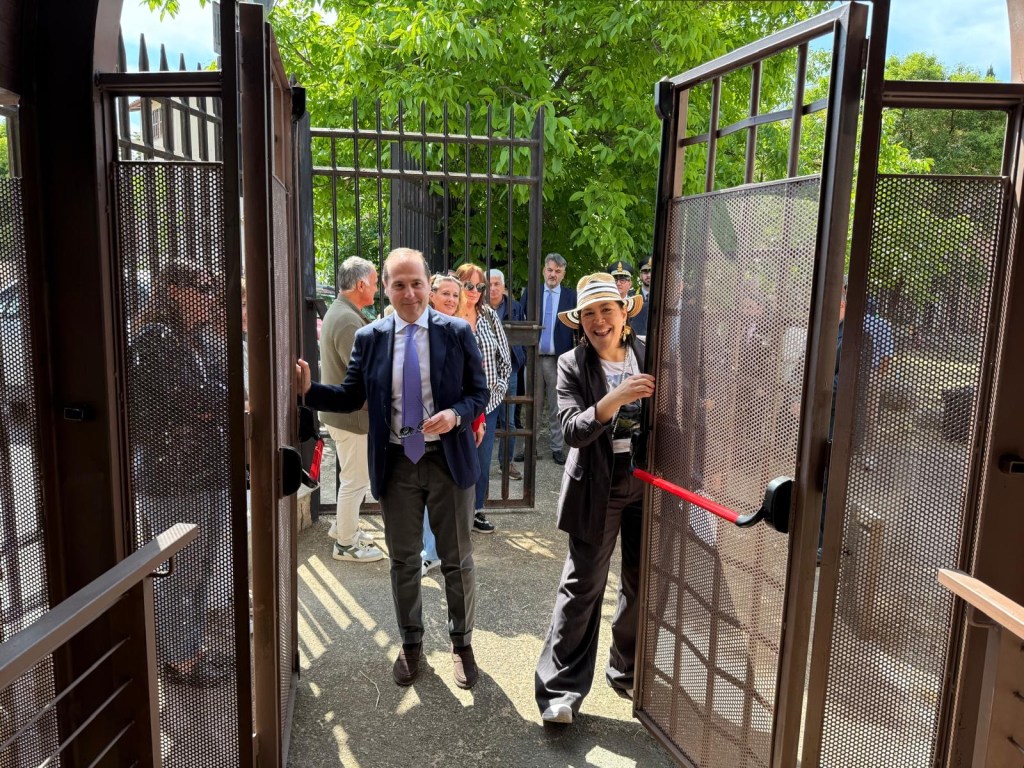 Two individuals, a man in a suit and a woman in a hat, are joyfully opening large gates, welcoming a group of people into an outdoor area surrounded by greenery.