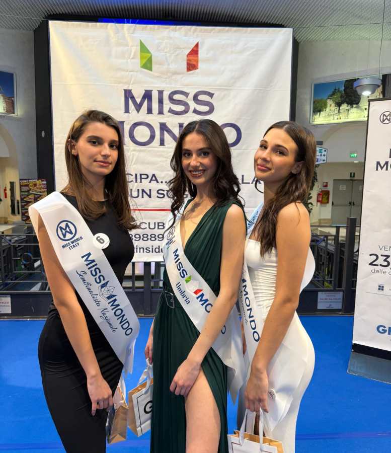 Tre finaliste di Miss Mondo Umbria 2025 posano insieme sul palco, indossando abiti eleganti e fasce con il logo del concorso, davanti a uno sfondo decorato con il logo di Miss Mondo.