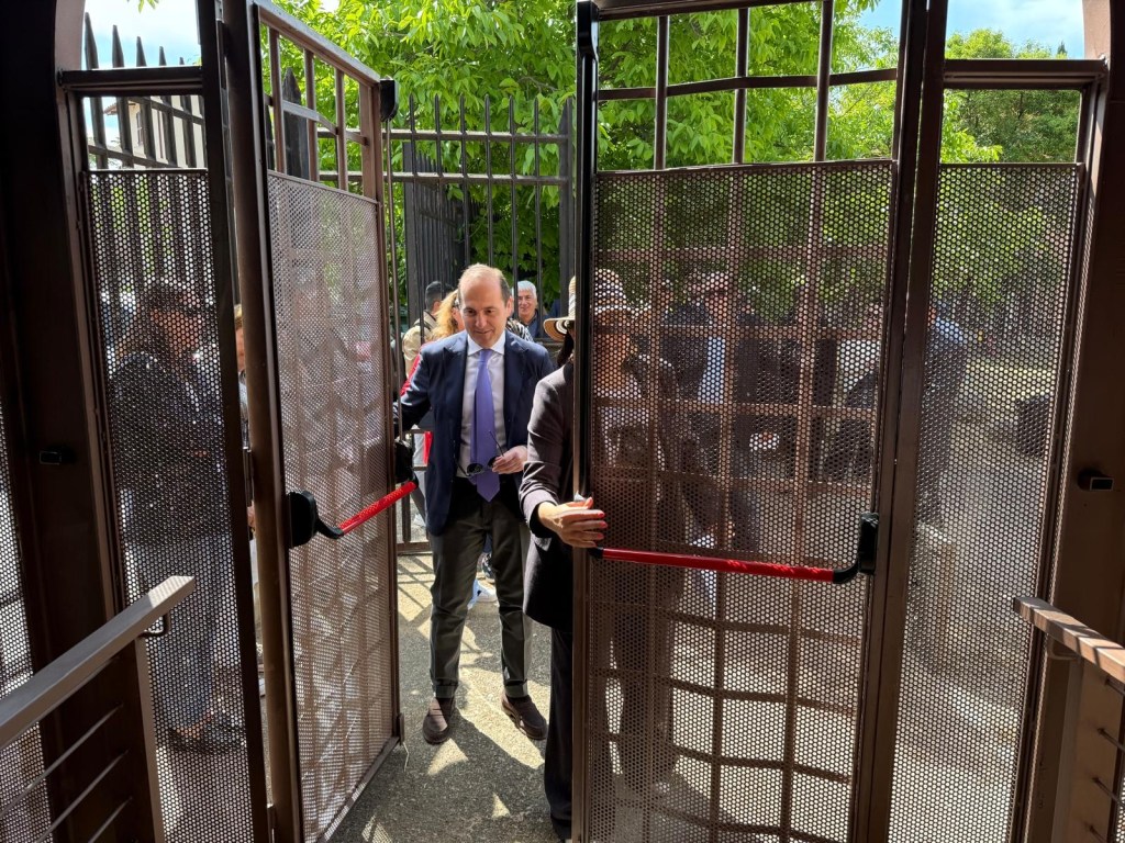 A group of people exiting through an open, gated entrance with mesh panels, with two individuals prominently pushing the gates. The scene captures a mix of greenery in the background and attendees dressed in business attire.