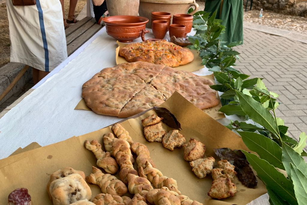 Un tavolo imbandito con una varietà di cibi antichi, tra cui focacce, dolci e pane, accompagnato da recipienti in terracotta e decorazioni vegetali per l'evento Ocriculum AD 168.