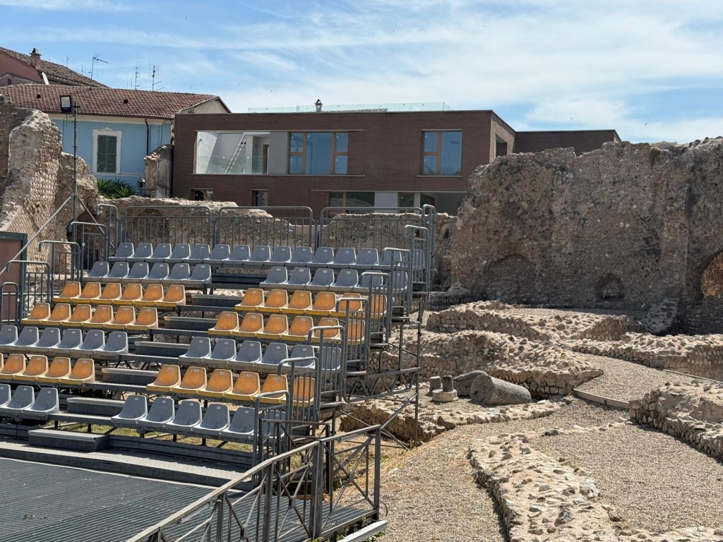 Ancient ruins with modern seating and a contemporary building in the background under a clear blue sky.