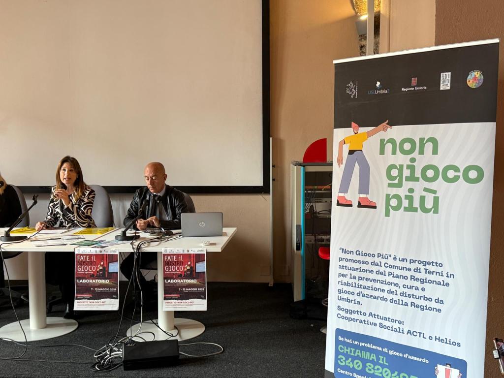 A meeting taking place with three speakers seated at a table, discussing the 'Non Gioco Più' initiative. In the background, a banner promoting the project is visible.