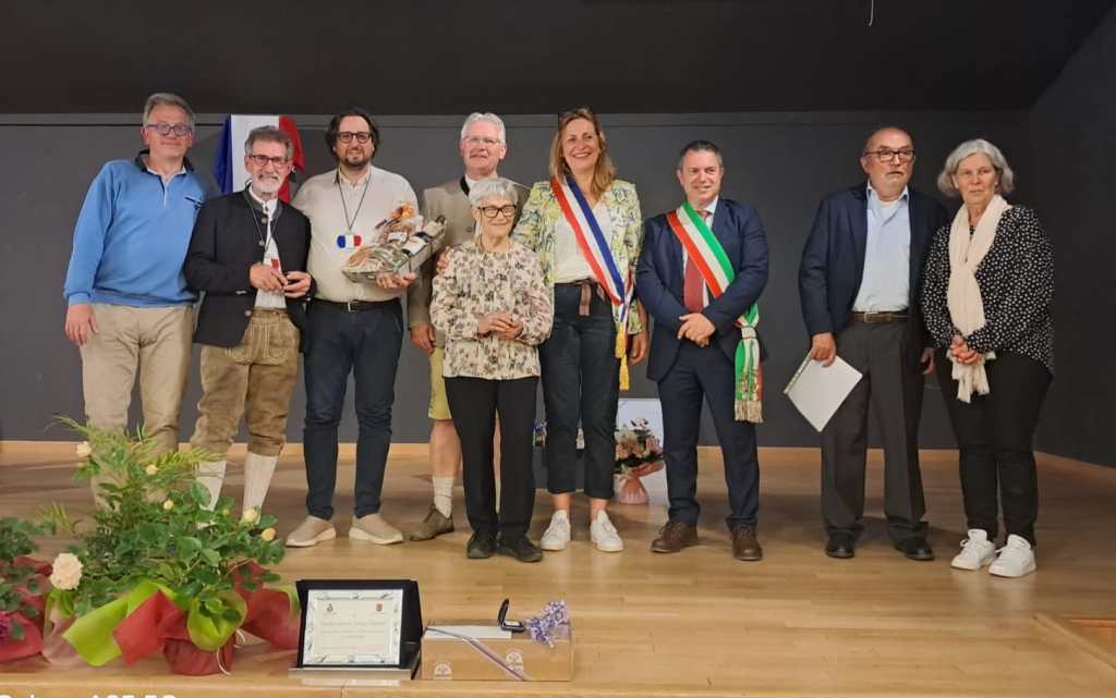 Un gruppo di persone sorridenti posano insieme sul palco, durante la celebrazione del 25° anniversario del gemellaggio tra Corciano e Civrieux d’Azergues. Alcuni membri indossano fasce tricolori e ci sono decorazioni floreali in primo piano.
