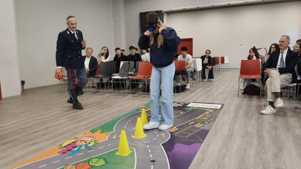Un giovane studente indossa un visore VR mentre cammina su un tappetino esperienziale, affiancato da un ufficiale di polizia. Sul tappetino sono presenti coni di traffico e un design che simula una strada, in un'aula con studenti seduti in background.