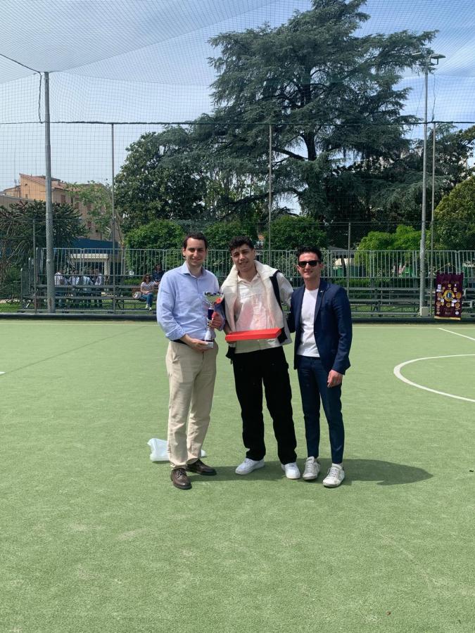Tre uomini sorridenti si incontrano su un campo da calcio, uno tiene un trofeo e un altro una teglia rossa.