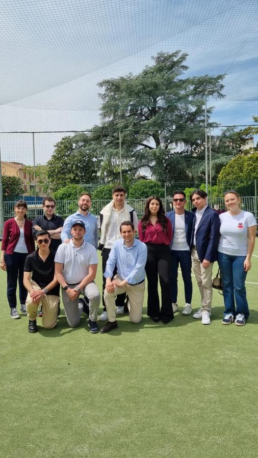 Gruppo di persone sorridenti in piedi e inginocchiati su un campo sportivo, con alberi e rete sullo sfondo.