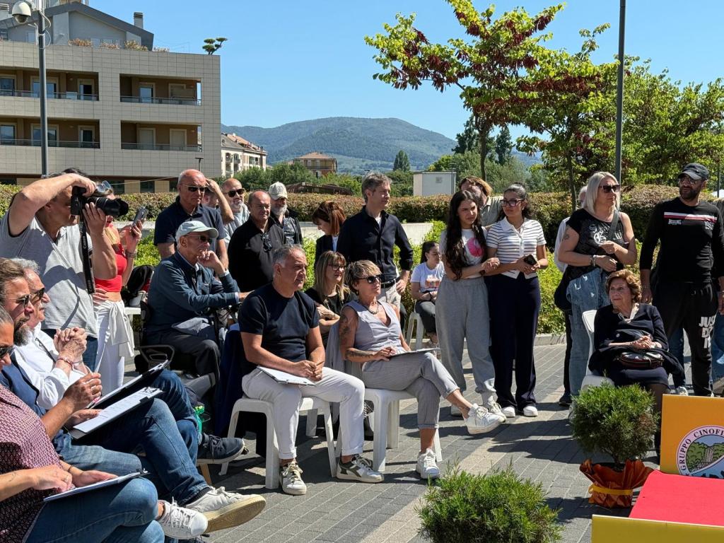 A gathering of people outdoors, some seated while others stand, with a backdrop of buildings and trees under a clear blue sky.