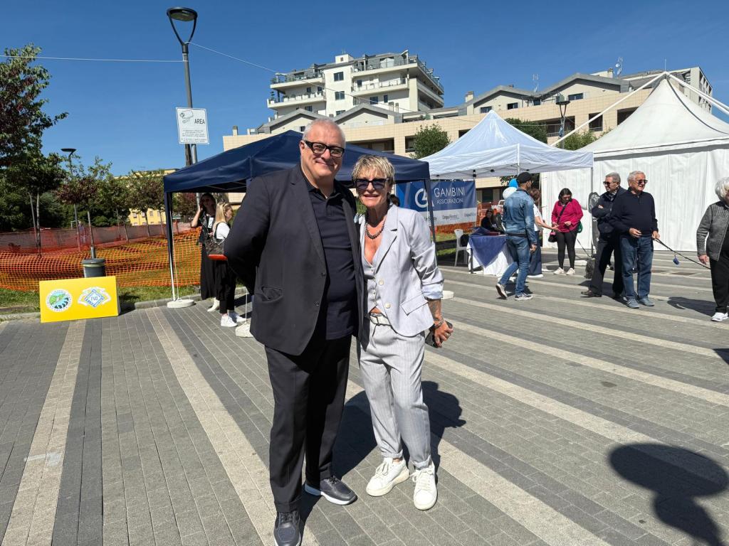 Due persone sorridenti in un evento all'aperto, circondate da stand e visitatori.