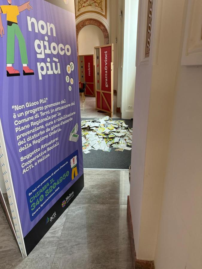 A colorful banner with the text 'non gioco più' stands next to an open doorway leading to a room with scattered papers on the floor.