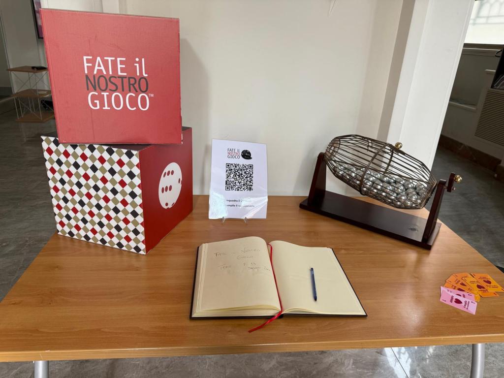 A wooden table displaying a lottery setup, featuring two decorative boxes with 'FATE il NOSTRO GIOCO' branding, a bingo cage filled with balls, a guest book open with a pen, and colored tickets laid out beside it.