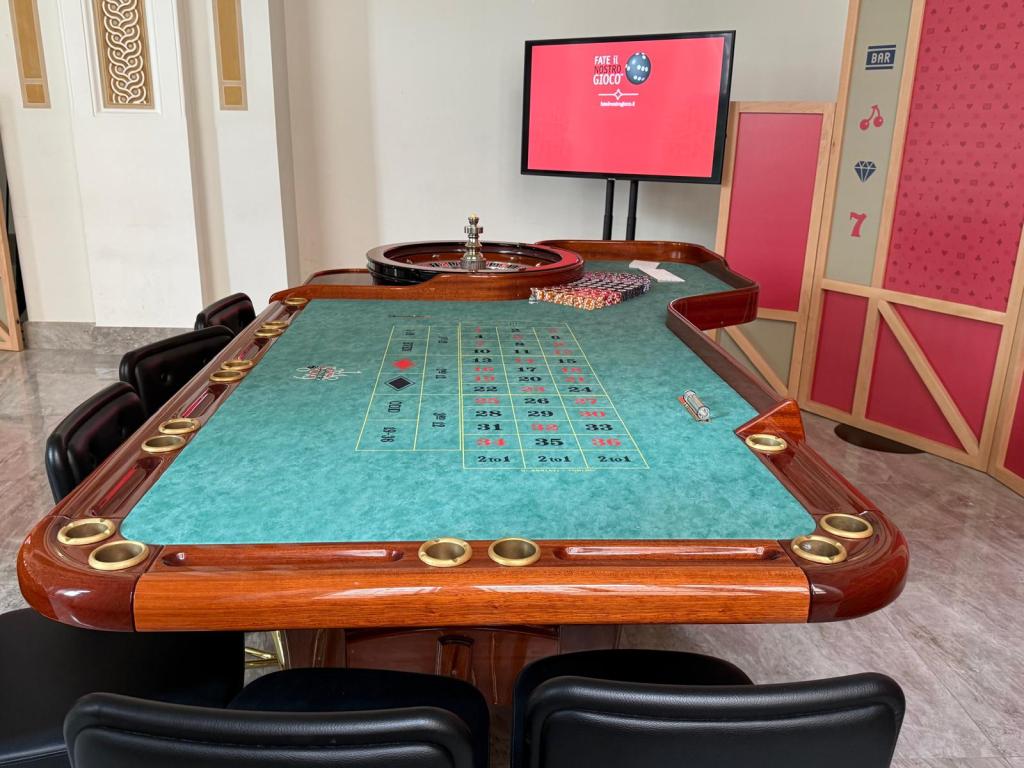 A roulette table with a green felt surface, surrounded by black chairs. A digital screen displaying text is in the background, along with chips and a roulette wheel on the table.