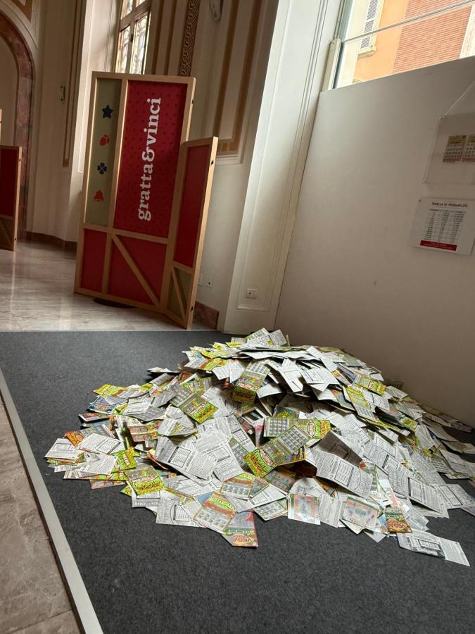 A large pile of scratch-off lottery tickets on a gray surface, with a backdrop featuring a display board labeled 'gratta&vinci'.
