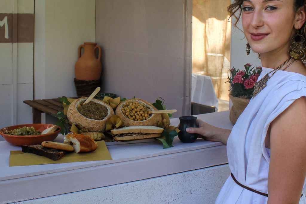 A woman in a white dress holds a black cup while standing near a table with traditional foods, including bowls of lentils and chickpeas, bread, and pastries, with decorative flowers in the background.