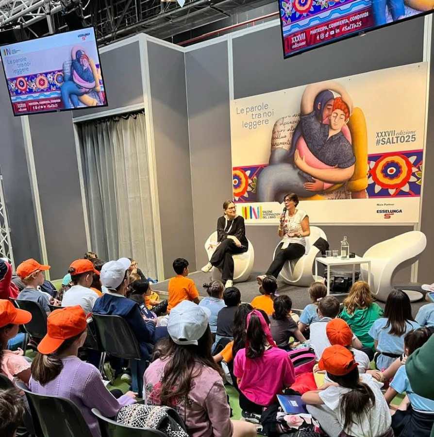 Un gruppo di bambini ascolta un incontro con l'autrice Patrizia Fortunati al Salone Internazionale del Libro di Torino, con due relatori sul palco e un grande poster sullo sfondo.