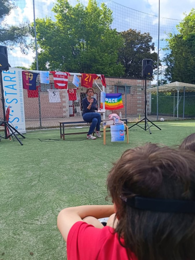 Un'autrice in una presentazione del libro 'Noi siamo la Belinda!' su un campo sportivo, circondata da bandiere colorate e magliette appese, mentre i bambini ascoltano attentamente.