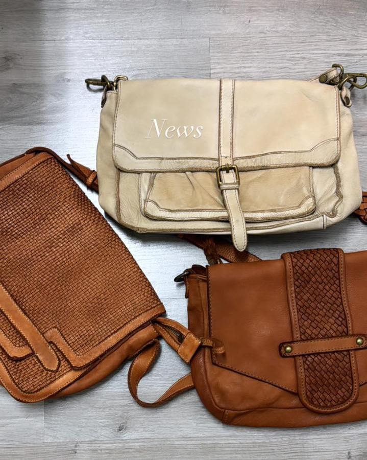 Tre borse in pelle di diversi colori e stili, disposte su un tavolo in legno chiaro. La borsa centrale è chiara con la scritta 'News', mentre le altre due sono di tonalità marrone, una con finitura intrecciata.
