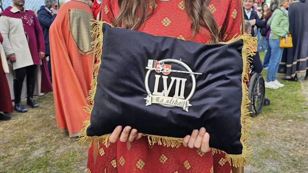 Una donna in abito tradizionale rossa tiene un cuscino nero con frange dorate e un simbolo in metallo che riporta il numero LVII e la scritta 'La stiba'.