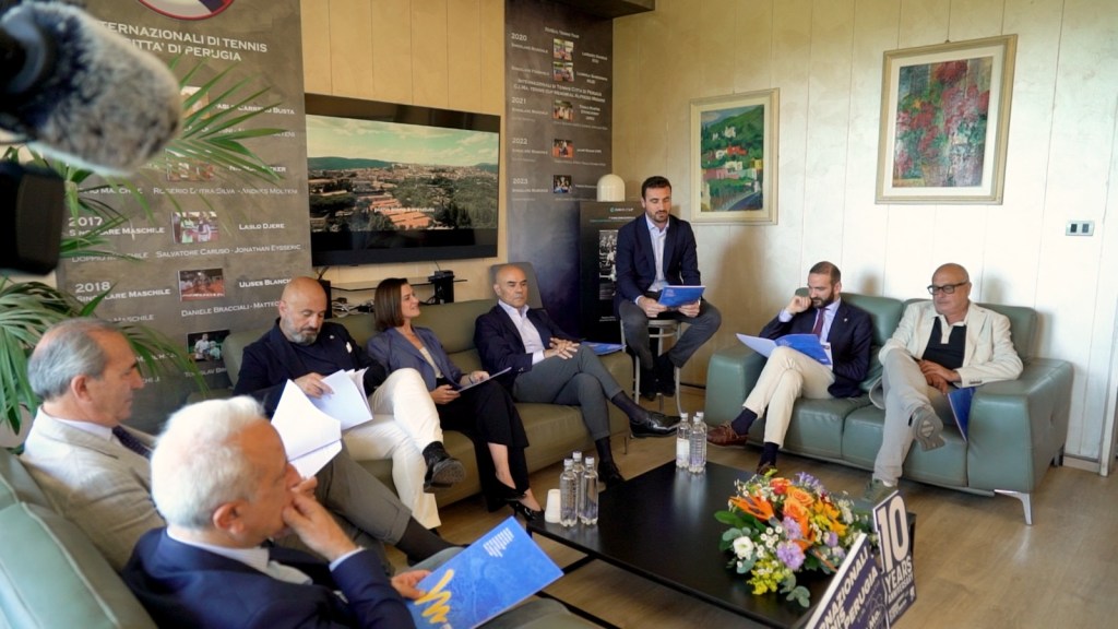Una conferenza stampa per presentare gli Internazionali di Tennis Città di Perugia, con vari relatori seduti su un divano e un tavolo decorato con fiori, bottiglie d'acqua e materiale informativo.