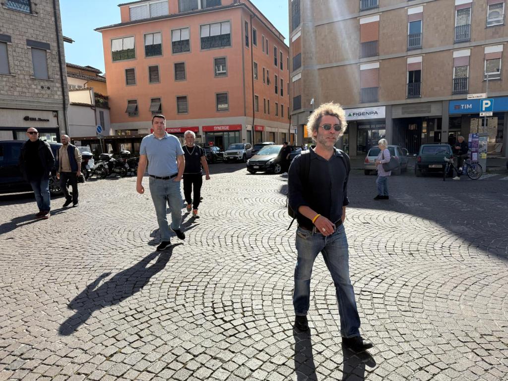 A bustling urban scene featuring several individuals walking on a cobblestone street under bright sunlight, with buildings in the background.