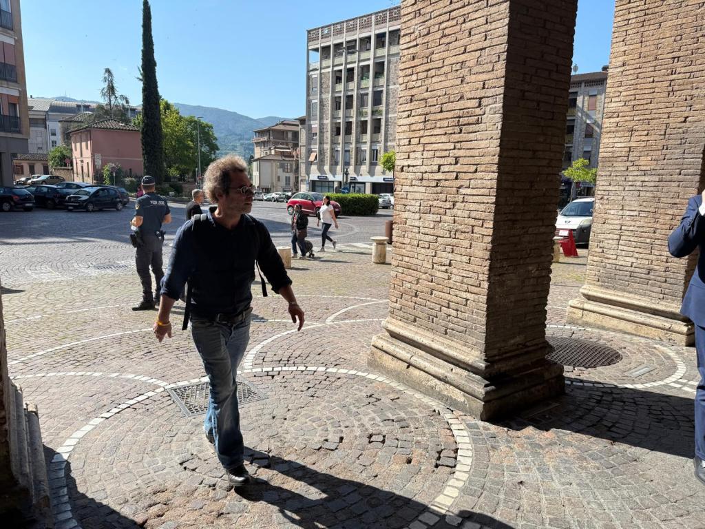 A person walking in a plaza with brick columns, surrounded by parked cars and buildings in the background on a sunny day.