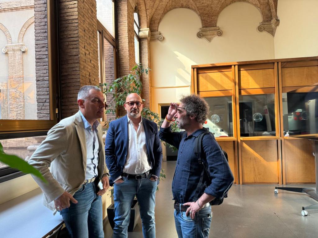 Three men are engaged in a conversation in a well-lit room with large windows, featuring brick walls and wooden furnishings.