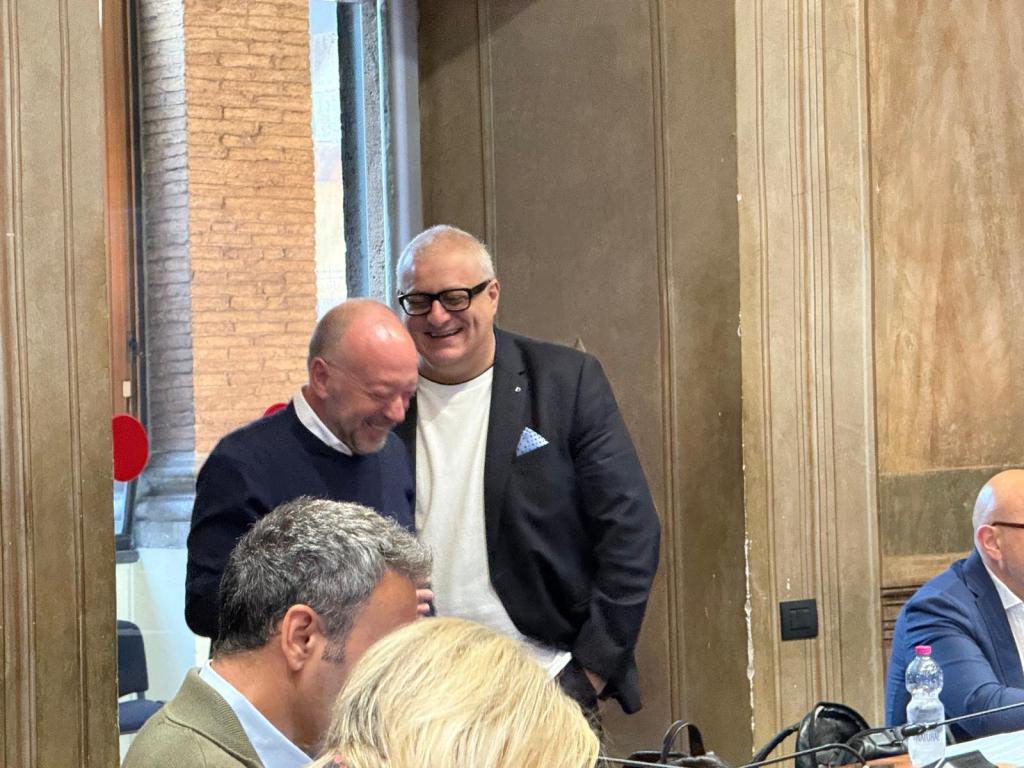 Two men laughing and engaging in conversation in a formal setting, with wooden paneling and a large window in the background.