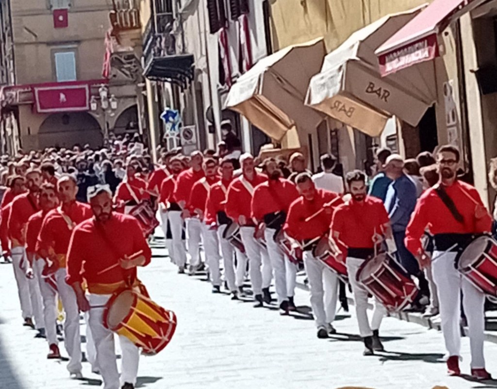 Parata di batteristi in maglietta rossa e pantaloni bianchi durante un evento tradizionale a Gubbio, con una folla che osserva lungo la strada.