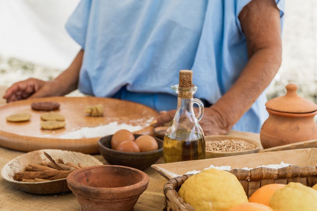 Un tavolo con ingredienti tradizionali romani, tra cui uova, olio d'oliva, limoni e erbe, mentre una persona con una tunica blu prepara pasticcini su un piano di legno.