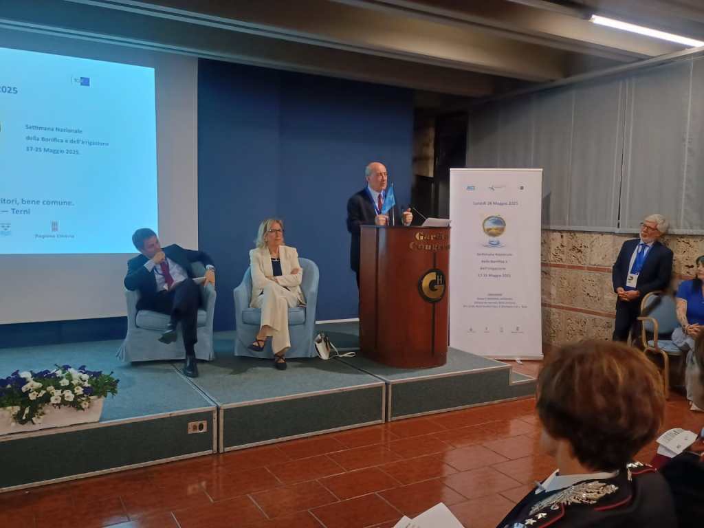 A conference scene with a speaker presenting at a podium, flanked by seated audience members. A banner with event details is visible in the background.
