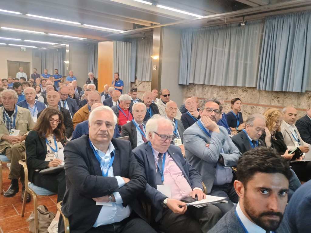 A large audience seated in a conference room, attentively listening to a speaker with a mix of older and middle-aged individuals.