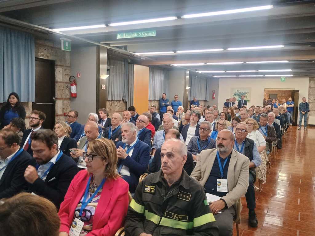 A crowded conference room with attendees listening attentively, featuring a diverse group of people wearing name tags and seated in rows.